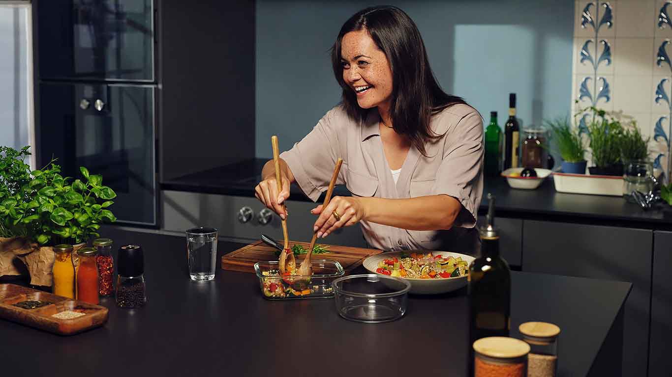 Une femme, l'air heureux, est dans sa cuisine en train de pr&eacute;parer un plat sain et color&eacute;.